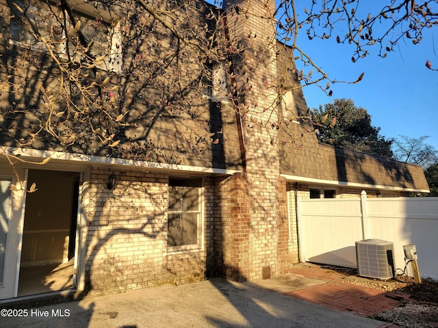 view of side of home featuring fence, brick siding, central AC, and a chimney