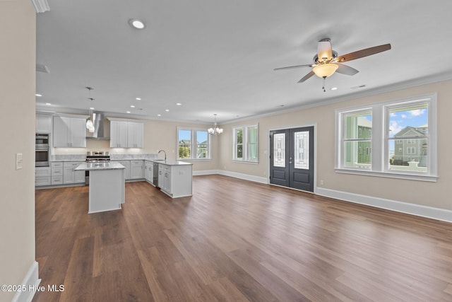 kitchen with wall chimney range hood, pendant lighting, sink, an island with sink, and ceiling fan with notable chandelier