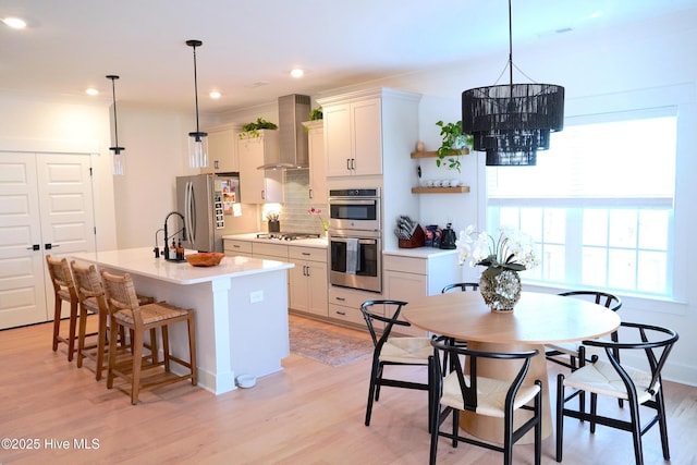 kitchen with light hardwood / wood-style floors, a center island with sink, appliances with stainless steel finishes, decorative light fixtures, and wall chimney range hood