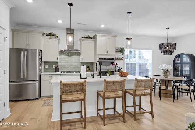 kitchen with stainless steel appliances, hanging light fixtures, wall chimney range hood, and a center island with sink