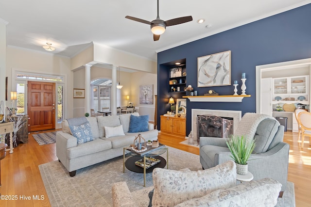 living room with built in shelves, ornate columns, light hardwood / wood-style floors, ornamental molding, and ceiling fan