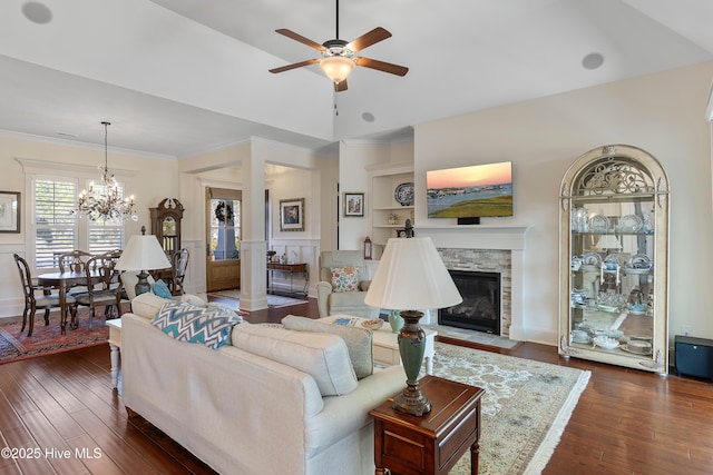 living room with dark wood-type flooring, a stone fireplace, crown molding, built in features, and ceiling fan with notable chandelier