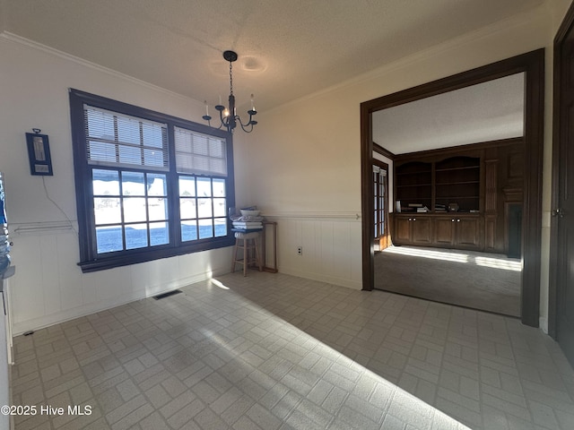 unfurnished dining area featuring a textured ceiling, a notable chandelier, a wainscoted wall, visible vents, and crown molding