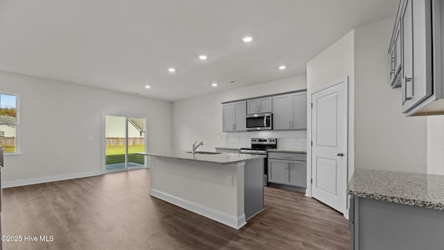 kitchen with light stone counters, stainless steel appliances, sink, gray cabinets, and an island with sink