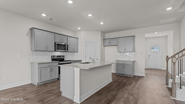 kitchen with light stone countertops, stainless steel appliances, dark hardwood / wood-style flooring, gray cabinets, and a center island with sink