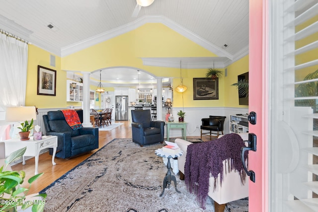 living area with arched walkways, a wainscoted wall, wood finished floors, vaulted ceiling, and ornate columns