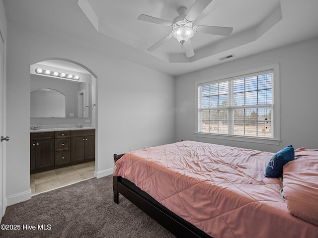 bedroom featuring ceiling fan, light colored carpet, connected bathroom, and a tray ceiling