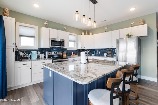 kitchen with pendant lighting, appliances with stainless steel finishes, a kitchen island with sink, backsplash, and white cabinetry