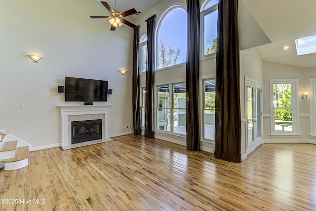 unfurnished living room with light wood-type flooring, ceiling fan, a skylight, and a high ceiling