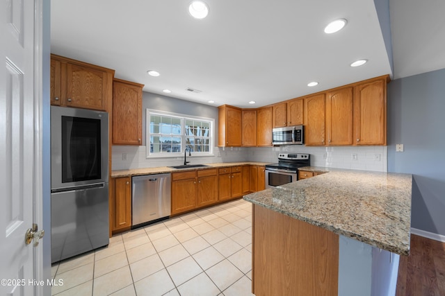 kitchen featuring appliances with stainless steel finishes, a sink, a peninsula, brown cabinetry, and light stone countertops