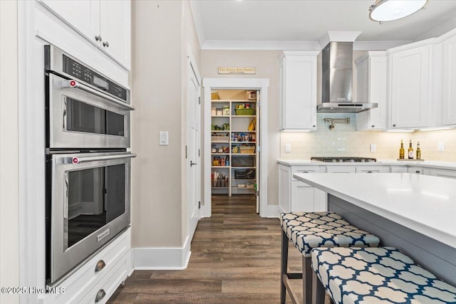 kitchen with double oven, gas stovetop, white cabinetry, light countertops, and wall chimney exhaust hood