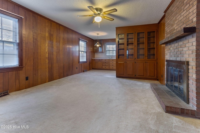 unfurnished living room featuring a textured ceiling, ceiling fan, wood walls, a brick fireplace, and light colored carpet