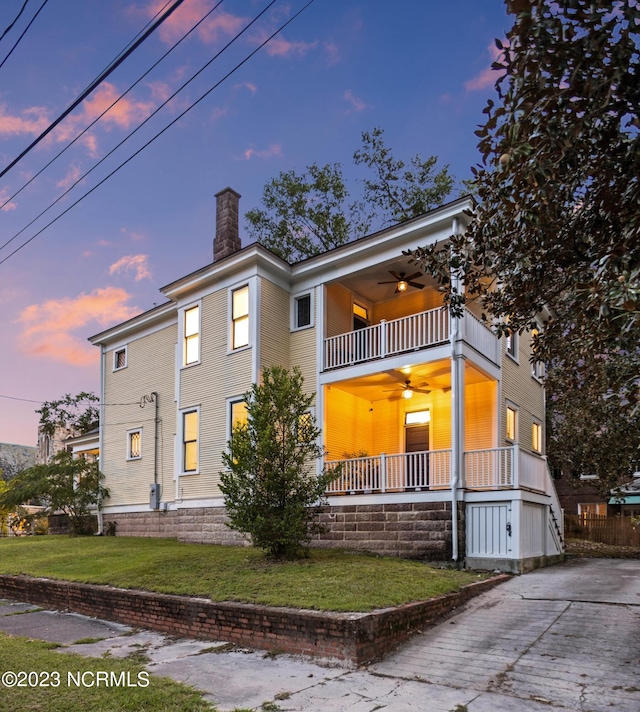 view of front of house with a balcony, a chimney, a ceiling fan, concrete driveway, and a yard