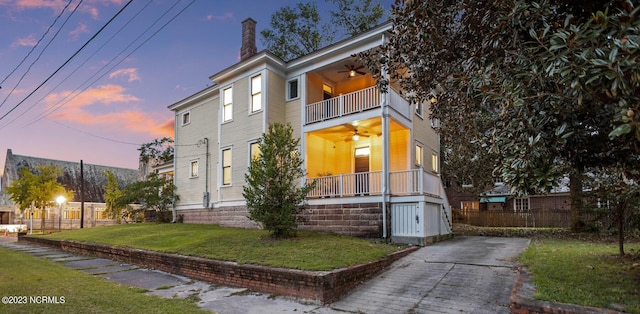 view of front facade with fence, a balcony, a chimney, a ceiling fan, and a lawn