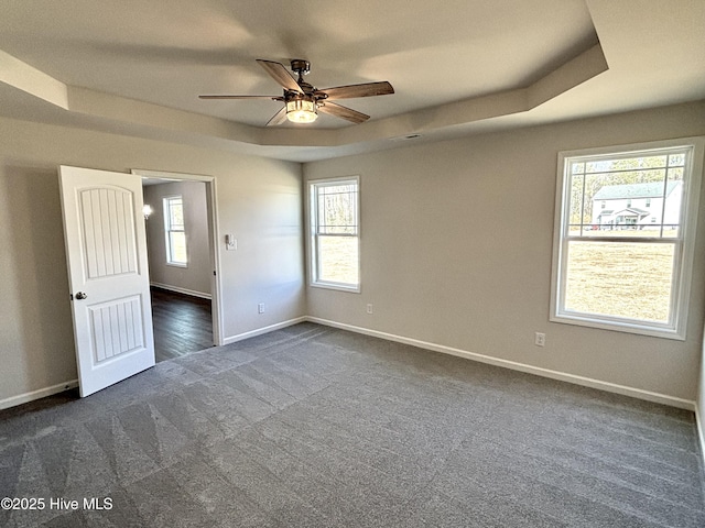 empty room with ceiling fan, a healthy amount of sunlight, dark carpet, and a tray ceiling