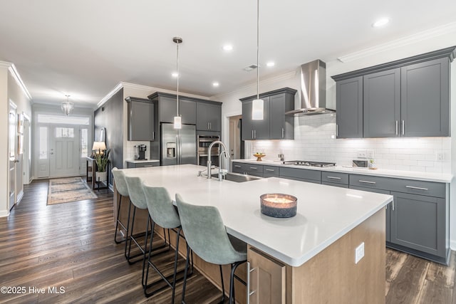 kitchen featuring pendant lighting, wall chimney exhaust hood, sink, a kitchen island with sink, and stainless steel appliances