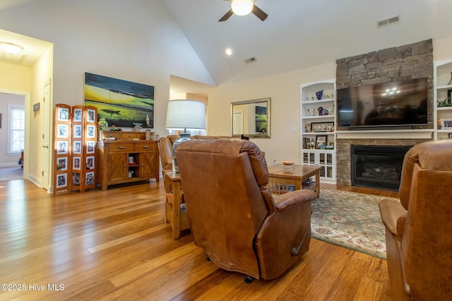 living room with a stone fireplace, high vaulted ceiling, ceiling fan, built in shelves, and light hardwood / wood-style flooring