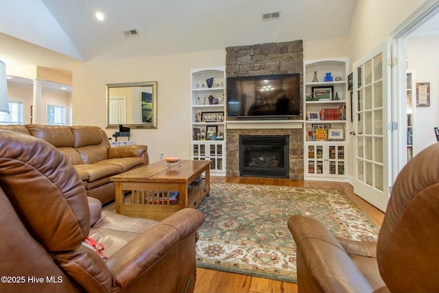 living room with ornate columns, vaulted ceiling, a stone fireplace, wood-type flooring, and built in shelves