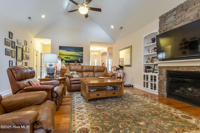 living room with hardwood / wood-style flooring, ceiling fan with notable chandelier, high vaulted ceiling, built in shelves, and a stone fireplace