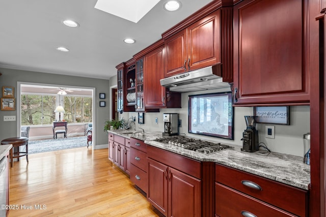 kitchen with a skylight, stainless steel gas stovetop, dark brown cabinets, light stone countertops, and under cabinet range hood