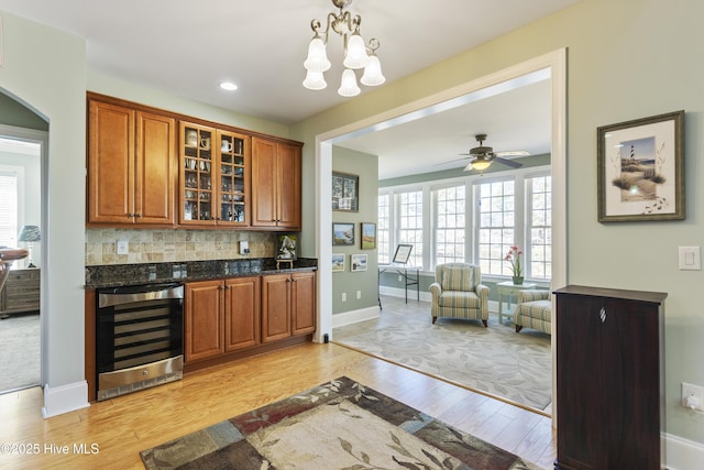 bar featuring backsplash, dark stone countertops, beverage cooler, ceiling fan with notable chandelier, and light hardwood / wood-style flooring