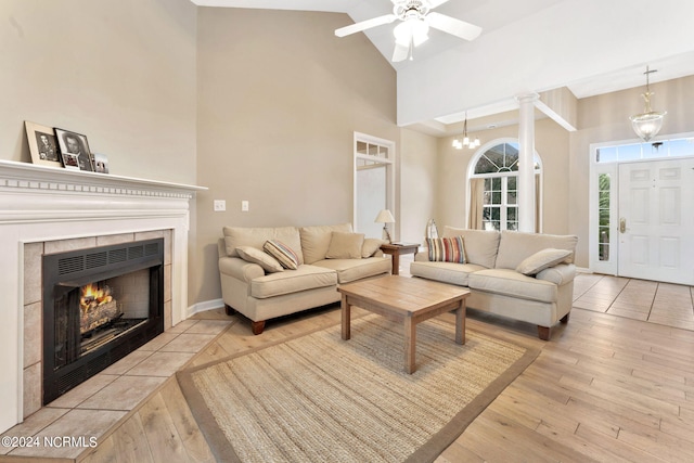 living room with a high ceiling, light wood-type flooring, ceiling fan, a tile fireplace, and decorative columns