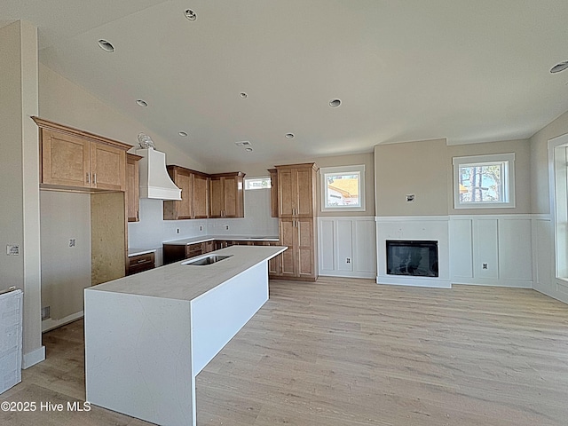 kitchen featuring light wood finished floors, a center island with sink, a glass covered fireplace, lofted ceiling, and custom range hood