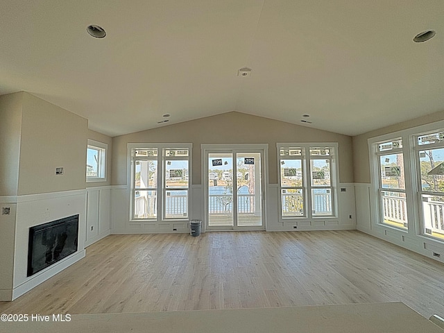 unfurnished living room featuring lofted ceiling, wainscoting, light wood finished floors, and a glass covered fireplace