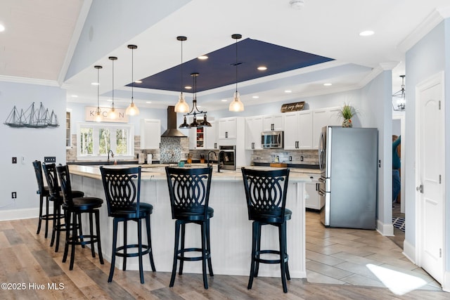 kitchen featuring stainless steel appliances, a tray ceiling, white cabinets, decorative light fixtures, and wall chimney exhaust hood