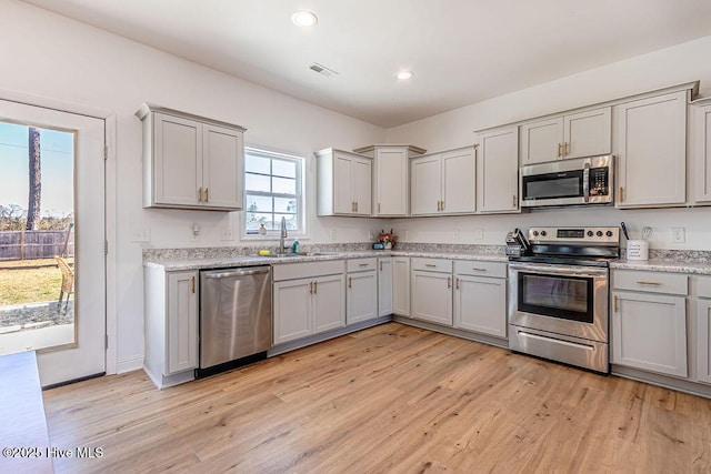 kitchen with gray cabinets, appliances with stainless steel finishes, sink, light stone countertops, and light hardwood / wood-style flooring