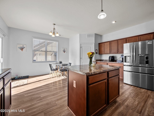 kitchen with stainless steel refrigerator with ice dispenser, hanging light fixtures, dark hardwood / wood-style flooring, a kitchen island, and dark stone counters