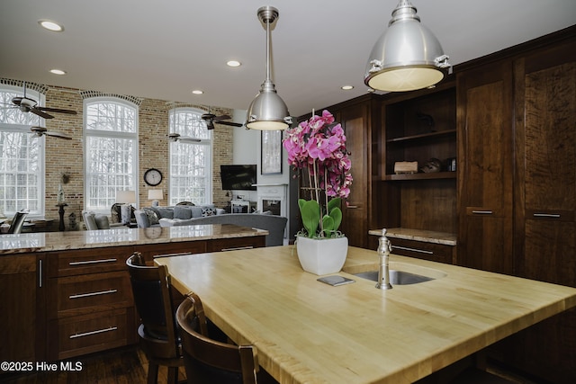 kitchen with ceiling fan, brick wall, a fireplace, dark brown cabinets, and light stone countertops