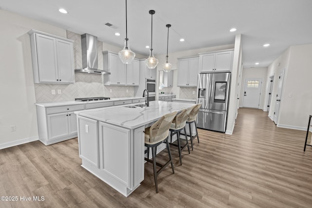 kitchen with white cabinets, wall chimney exhaust hood, stainless steel appliances, a kitchen island with sink, and light stone counters