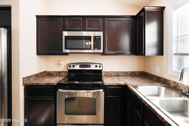 kitchen with lofted ceiling, a sink, dark brown cabinets, appliances with stainless steel finishes, and dark countertops