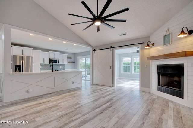 unfurnished living room with vaulted ceiling, a textured ceiling, ceiling fan, a barn door, and light hardwood / wood-style floors