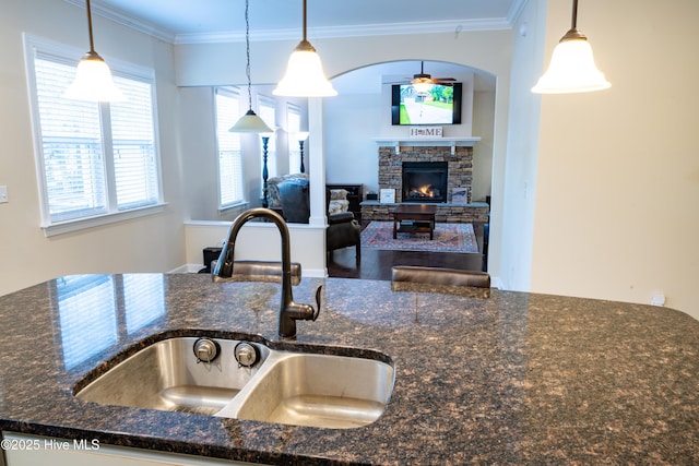 kitchen featuring a stone fireplace, sink, crown molding, decorative light fixtures, and dark stone counters