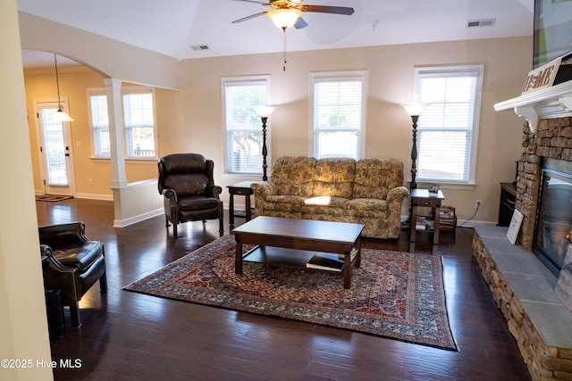 living room featuring dark wood-type flooring, a stone fireplace, ornate columns, vaulted ceiling, and ceiling fan