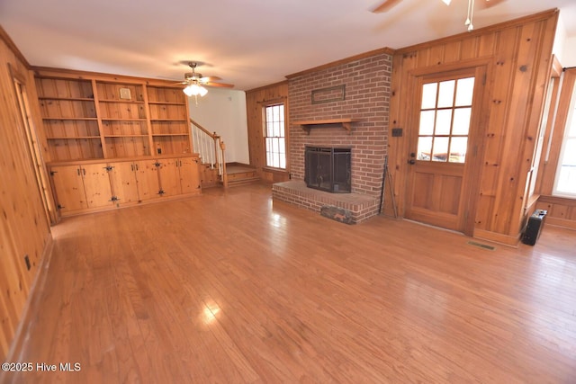 unfurnished living room with a brick fireplace, wooden walls, light wood-type flooring, built in shelves, and ceiling fan