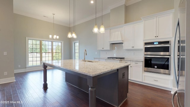 kitchen featuring light stone countertops, white cabinetry, premium range hood, hanging light fixtures, and a kitchen island with sink