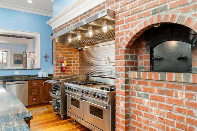 kitchen featuring brown cabinetry, light wood finished floors, ornamental molding, stainless steel appliances, and wall chimney range hood