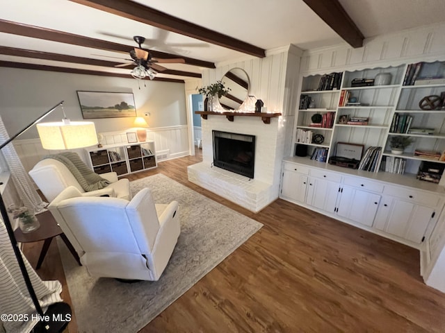 living room featuring a brick fireplace, dark wood-type flooring, beamed ceiling, and ceiling fan