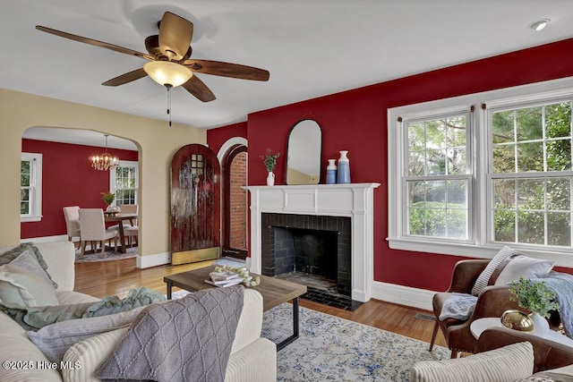 living area with arched walkways, a brick fireplace, wood finished floors, baseboards, and ceiling fan with notable chandelier