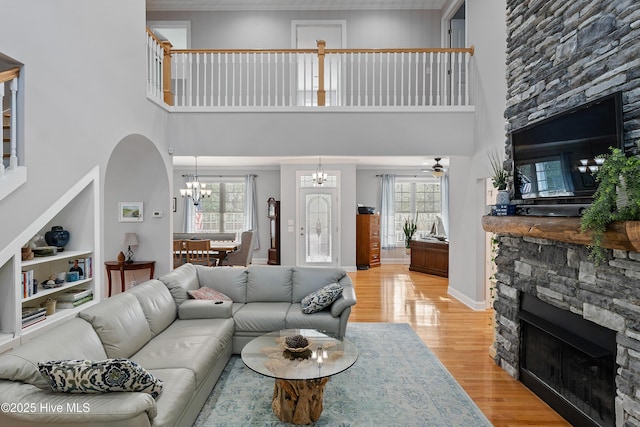 living room featuring arched walkways, a stone fireplace, light wood-type flooring, baseboards, and ceiling fan with notable chandelier