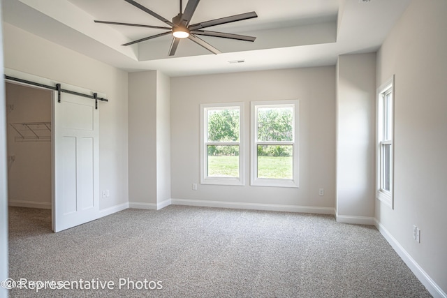 unfurnished bedroom with a walk in closet, light colored carpet, a tray ceiling, ceiling fan, and a barn door