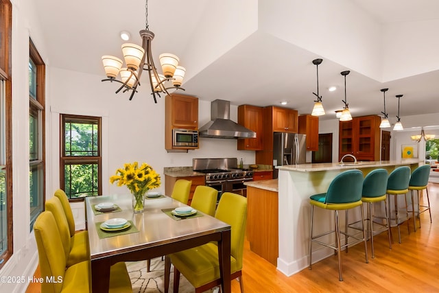 kitchen with appliances with stainless steel finishes, pendant lighting, a chandelier, wall chimney exhaust hood, and light hardwood / wood-style flooring