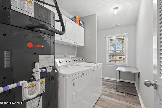 clothes washing area with washing machine and clothes dryer, heating unit, cabinets, light hardwood / wood-style flooring, and a textured ceiling