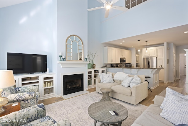 living room featuring a towering ceiling, light hardwood / wood-style flooring, a tile fireplace, and ceiling fan