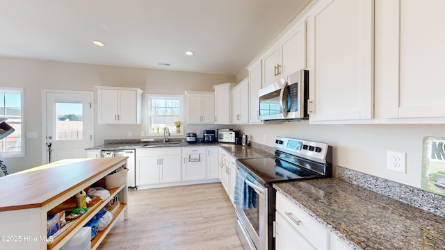 kitchen with white cabinets, stainless steel appliances, dark stone countertops, a sink, and light wood-type flooring