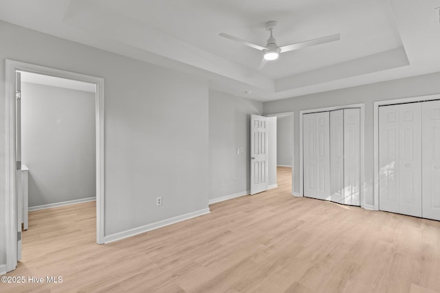 unfurnished bedroom featuring multiple closets, ceiling fan, a tray ceiling, and light hardwood / wood-style floors