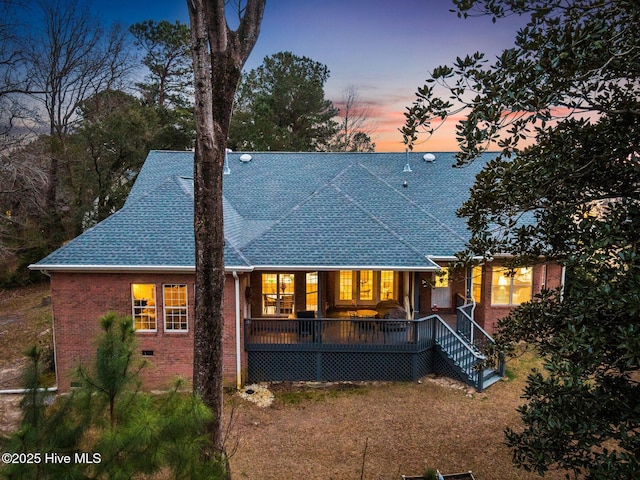 back of property at dusk with crawl space, a wooden deck, brick siding, and a shingled roof